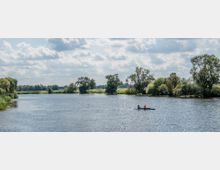 Flusslandschaft mit breitem Wasserlauf, gesäumt von grünen Bäumen und Wiesen; im Vordergrund ein Zweier-Kajak mit zwei Personen. Der Himmel ist teils bewölkt, und das Wasser glitzert im Sonnenlicht.
