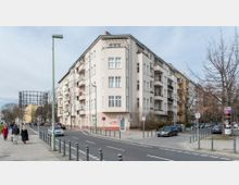Eckgebäude eines Wohnhauses in einer städtischen Straße mit hellen Fassaden und Balkonen, umgeben von Gehwegen, parkenden Autos und kahlen Bäumen. Im Hintergrund ist ein großer Gasometer zu sehen, während Fußgänger am Straßenrand unterwegs sind.