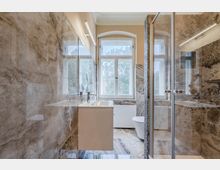 Modernes, schmales Badezimmer mit großformatigen marmorierten Wand- und Bodenfliesen, Waschtisch und Spiegel an der linken Wand sowie einer verglasten Dusche rechts. Am Ende des Raums befindet sich ein großes Fenster mit Oberlicht über einem Heizkörper, daneben ein WC.