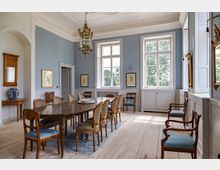 Eleganter Speiseraum mit hellblauen Wänden, Parkettboden und einem langen Holztisch umgeben von gepolsterten Stühlen. Große Fenster lassen natürliches Licht herein, und die Einrichtung umfasst goldgerahmte Bilder, einen Kronleuchter und eine klassische Konsole mit einer Vase.