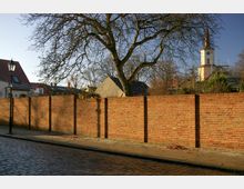Straßenszene mit einer roten Backsteinmauer entlang eines Bürgersteigs, dahinter ein kahler Baum und mehrere Gebäude, darunter ein Kirchturm mit Uhr und spitzem Dach. Der Vordergrund zeigt Kopfsteinpflaster, eine Laterne steht links neben der Mauer.