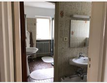 Badezimmer mit beigen, gemusterten Fliesen an den Wänden, einem Waschbecken mit Wandspiegel, einem Heizkörper unter einem Fenster und braunen Fliesen auf dem Boden. Auf dem Boden liegen mehrere helle Teppiche.