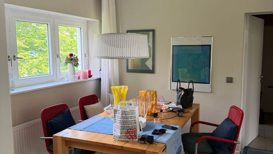 Esszimmer mit rechteckigem Holztisch, blauem Tischläufer und vier rot gepolsterten Stühlen; darüber hängt eine große, zylindrische Pendelleuchte. Links befindet sich ein Doppelfenster mit weißen Vorhängen und Blick ins Grüne, an der Wand hängen gerahmte Bilder und rechts führt eine offene Tür in einen angrenzenden Flur.
