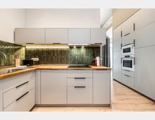 Moderne Einbauküche mit weißen, grifflosen Schränken, Holzarbeitsplatte und grün glasiertem Fliesenspiegel mit Unterbauleuchten. Rechts befindet sich ein hoher Schrankblock mit integriertem Backofen und Mikrowelle, der Raum hat hellen Holzboden und eine schlichte, helle Wandgestaltung.