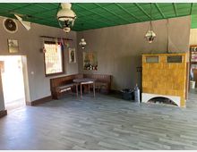 Ein rustikaler Innenraum mit grauem Laminatboden, grüner Decke und hellbraunen Wänden. Links steht eine Eckbank mit Esstisch, rechts ein gelber Keramikofen mit Verzierungen; an der Decke hängen mehrere alte Lampen.