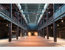 Großer, modern gestalteter Innenraum mit industriellem Charakter, geprägt von schwarzen Stahlträgern, oberen Galerien und Bühnenbeleuchtung an der Decke. Der Boden ist glatt, und am Ende des Raums befindet sich eine Ziegelwand mit einer Tür in der Mitte.