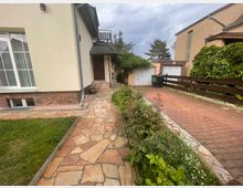 Einfamilienhaus mit heller Fassade, rotem Klinkersockel und Schieferschindeldach; gepflasterter Weg führt entlang eines kleinen Gartens mit Blumen und Sträuchern zu einer überdachten Eingangstür. Rechts im Bild eine gepflasterte Einfahrt mit einer Garage, Holzzaun und bewachsener Hecke.