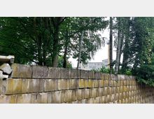Eine niedrige Mauer aus Betonblöcken im Vordergrund, dahinter Bäume mit dichtem Laub. Im Hintergrund sind Teile eines mehrstöckigen Gebäudes und ein hoher Schornstein zu sehen.