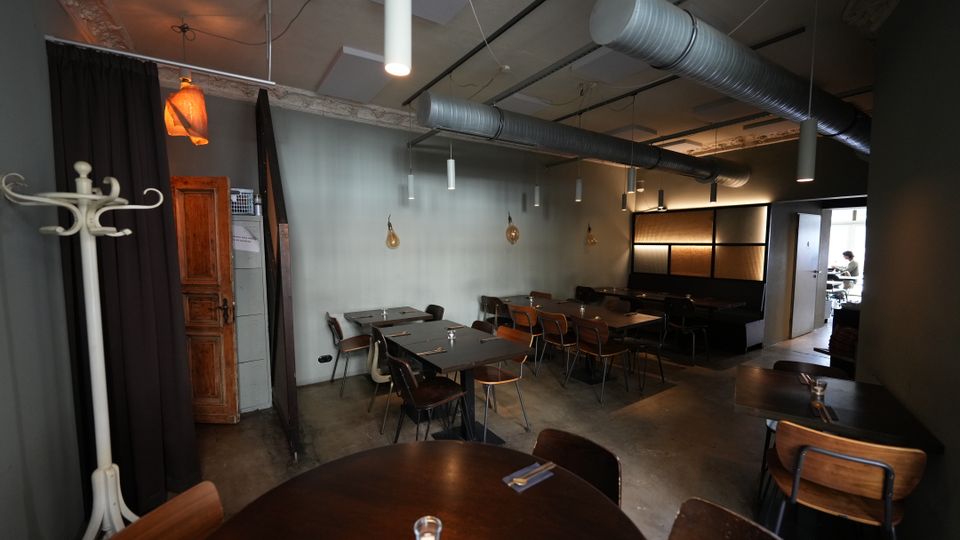 Innenraum eines modernen Restaurants mit mehreren dunklen Holztischen und Stühlen auf Betonboden. Sichtbare Lüftungsrohre und schlichte Hängelampen an der Decke sowie eine Sitzecke mit gepolsterter Bank und abgetrenntem Bereich im Hintergrund.