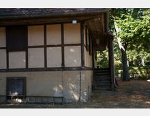 Fachwerkhaus mit hellen Putzwänden und dunklen Holzstreben, seitlich befindet sich eine hölzerne Treppe, die zu einem überdachten Eingang führt. Im Hintergrund sind Bäume und ein Zaun zu sehen.