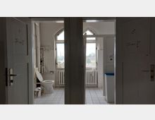 Zweifacher Sanitärraum mit weißen Fliesen, getrennt durch eine Wand, jeweils mit beschrifteten Holztüren (Symbol für Frauen auf der linken, für Männer auf der rechten Tür). Im Hintergrund zwei Fenster mit Spitzbogen oben, alte Heizkörper darunter, sowie eine Toilette und Mülleimer links und ein Papierkorb rechts.