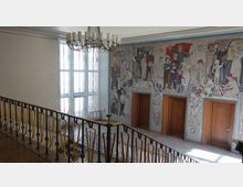 Treppenhaus eines Gebäudes mit einem farbenfrohen Wandmosaik, das Szenen von Menschen bei verschiedenen Tätigkeiten zeigt. Im Vordergrund ein Geländer mit goldfarbenen Details, im Hintergrund große Fenster mit geteilten Scheiben und drei Holztüren in der Wand mit dem Mosaik.