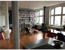 Offener Wohnraum mit Holzboden, hohen verzierten Säulen und großen Bogenfenstern, die viel Tageslicht hereinlassen. Im Raum befinden sich Regale voller Bücher, ein Kaminofen, ein Esstisch mit Stühlen und verschiedene Sitzmöbel.