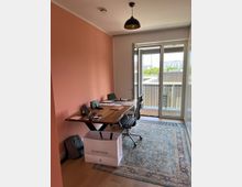 Helles Büro mit einer korallenfarbenen Wand, einem Holztisch mit technischen Geräten und einem schwarzen Bürostuhl auf einem orientalischen Teppich. Im Hintergrund befinden sich große Glastüren, die auf einen Balkon mit Blick auf moderne Gebäude führen.