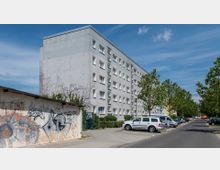 Ein mehrstöckiges Wohngebäude aus Beton mit kleinen rechteckigen Fenstern, daneben ein Parkplatz mit mehreren Autos. Im Vordergrund ist eine mit Graffiti besprühte Mauer, und die Straße ist von Bäumen gesäumt.