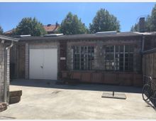 Hinterhof mit einem Backsteingebäude im Industrie-Stil, das große Fenster mit Metallrahmen und eine weiße Metalltür aufweist. Im Vordergrund befindet sich ein betonierter Boden, ein abgestelltes Fahrrad und Schatten von umliegender Bebauung sowie Bäume im Hintergrund.