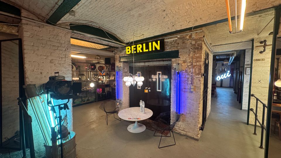 Industrie-inspirierter Innenraum mit weiß gestrichenen Backsteinwänden, sichtbaren Rohren und einer gelben Leuchtschrift mit der Aufschrift „BERLIN“. Im Vordergrund stehen ein runder Tisch und Stühle, umgeben von modernem Dekor und neonbeleuchteten Elementen.