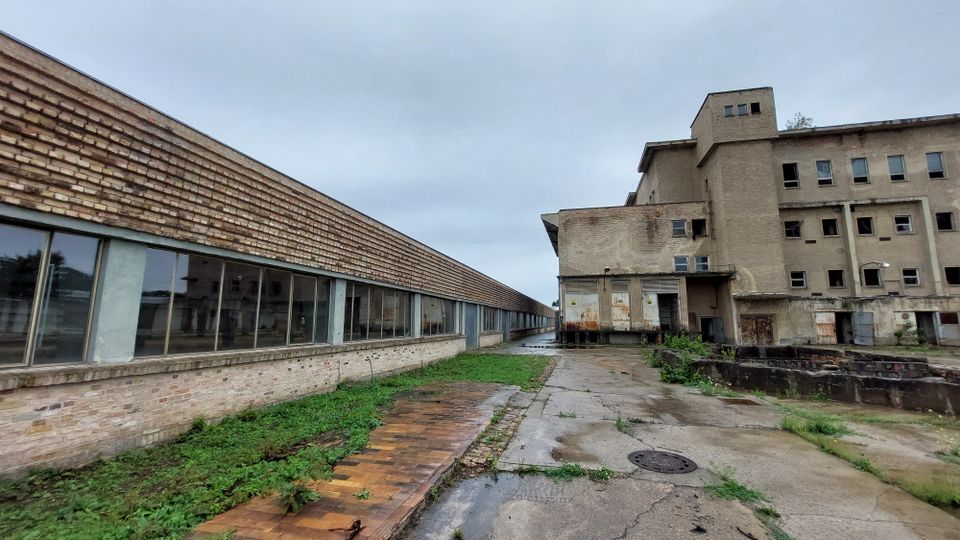 Ein verlassenes, teilweise verfallenes Industriegebäude mit Backsteinfassaden, großen Fensterfronten und rostigen Metalltüren. Der Boden besteht aus verwittertem Beton und Pflaster, teils überwuchert von Unkraut, unter wolkenbedecktem Himmel.