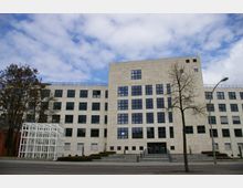 Mehrstöckiges Bürogebäude mit heller Fassade und großen, regelmäßigen Fenstern, davor eine breite Treppe mit Eingangstüren. Im Vordergrund stehen kahle Bäume und eine weiße, kubische Skulptur.