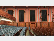 Innenraum eines Theatersaals mit Reihen von gepolsterten Stühlen in Blau. Die hölzernen Wände sind mit Türen und hohen, schwarzen Vorhängen gestaltet, die von pastellfarbenen Säulen in Rosa und Blau eingerahmt werden.