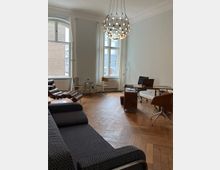 Heller Büroraum mit Fischgrätenparkettboden, großer Fensterfront mit weißen Vorhängen und einem dekorativen, kugelförmigen Kronleuchter an der Decke. Der Raum ist mit einem Schreibtisch aus Holz, braunen Stühlen und einer dunklen Liege ausgestattet.