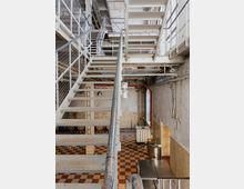 Innenansicht eines industriellen Gebäudes mit Metalltreppen, Gittergeländern und freiliegender Infrastruktur. Der Boden ist mit rot-beigen Fliesen ausgestattet, und im Hintergrund befinden sich Metallbehälter, Fässer und weitere industrielle Geräte.