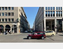 Straßenszene in einer Stadt mit zwei gegenüberliegenden Geschäftsgebäuden im modernen Stil, großen Fenstern und klaren Fassaden. Autos, Fahrräder und Fußgänger sind im Vordergrund auf einer Kreuzung zu sehen, darunter ein rotes Auto und ein Taxi; Straßenschilder und Geschäfte wie Douglas und Telekom sind erkennbar.