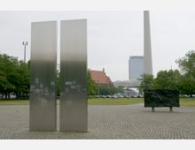 Zwei hohe, rechteckige Edelstahlsäulen mit eingelassenen Schwarz-Weiß-Bildern stehen auf einem gepflasterten Platz, umgeben von Rasenflächen und Bäumen. Im Hintergrund sind ein rotes Dach einer historischen Kirche, ein moderner Hochhausturm mit "Park Inn"-Logo und der Berliner Fernsehturm zu sehen.