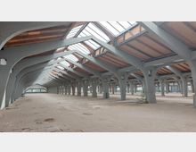 Weitläufiger, leerer Innenraum eines Gebäudes mit grauen Betonstreben, die das Holzdach mit Oberlichtern stützen. Der Boden besteht aus ungleichmäßigen Fliesen, und Licht fällt durch die großen Fenster am Ende des Raumes.