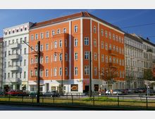 Straßenszene mit mehrstöckigem Wohnhaus in orangefarbenem Putz, flankiert von anderen hell verputzten Gebäuden; im Erdgeschoss befinden sich Geschäfte, davor geparkte Autos, Straßenbahngleise und eine Grünfläche.