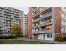 Wohnsiedlung mit mehrstöckigen Plattenbauten, deren Fassade teils aus Beton und teils mit braunen Kacheln verkleidet ist. Im Vordergrund befindet sich eine Grünfläche mit einem Baum und einem kleinen Spielplatz, eingerahmt von Büschen.