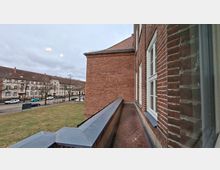Blick von einem schmalen Balkon an einem Backsteingebäude mit weißen Sprossenfenstern und rotbraunem Fliesenboden. Im Hintergrund sind eine Straße mit parkenden Autos, eine Rasenfläche und gegenüberliegende Wohnhäuser unter bewölktem Himmel zu sehen.