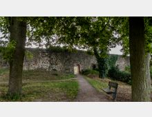 Ein mittelalterlicher Steinwall mit einem kleinen, gewölbten Durchgang aus Ziegelsteinen, umgeben von Bäumen und Grünflächen. Im Vordergrund steht eine Holzbank auf einer Wiese, und ein schmaler Weg führt zum Durchgang.