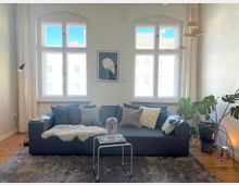 Helles Wohnzimmer mit einem großen, grauen Sofa, dekoriert mit Kissen und einer Decke, vor einem Fenster mit Rundbogenrahmen. Im Raum stehen eine Stehlampe, ein Beistelltisch und eine große Zimmerpflanze; Holzboden und minimalistische Einrichtung prägen den Stil.