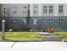 Hof eines Gebäudes mit grauer Fassade und weißen Fensterrahmen, umgeben von einer gepflegten Grünfläche mit Bänken, Treppenstufen und einer kugelförmigen Stein-Installation. Im Vordergrund ist ein gepflasterter Weg mit einem modernen Lampenmast zu sehen.