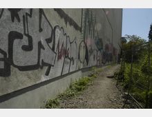 Eine lange Betonwand mit Graffiti, darunter bunte Schriftzüge und Zeichnungen, grenzt an einen schmalen, unbefestigten Weg mit Grasbewuchs. Rechts neben dem Weg steht ein metallener Zaun, und im Hintergrund befinden sich Bäume unter klarem Himmel.