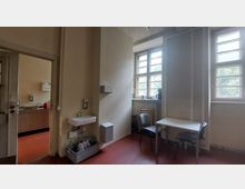 Ein einfacher Raum mit Waschbecken, einem kleinen Tisch und Stühlen, beleuchtet durch zwei große Fenster mit Holzrahmen. Im angrenzenden Bereich ist eine Küche mit Spülbereich und rotem Boden sichtbar.