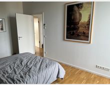 Schlafzimmer mit Parkettboden, einem Bett mit grauer Decke im Vordergrund, einer offenen weißen Tür, die zu einem angrenzenden Raum führt, sowie einem großformatigen gerahmten Foto an der weißen Wand.
