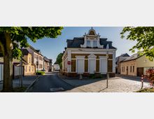 Straßenszene in einer kleinen Ortschaft mit einem historischen Gebäude an einer Straßengabelung, umgeben von Wohnhäusern und Bäumen. Das zentrale Gebäude hat Schmuckelemente an der Fassade, ein Mansardendach und geschlossene Fensterläden.