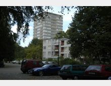 Ein Wohngebiet mit einem mehrstöckigen Hochhaus im Hintergrund und niedrigeren Wohngebäuden im Vordergrund, umgeben von Bäumen. Vor den Gebäuden stehen mehrere geparkte Autos auf einem gepflasterten Parkplatz.