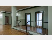 Flur eines Gebäudes mit holzfarbenem Parkettboden, grün gestrichenen Wänden und mehreren Türen mit Milchglas-Einsätzen und dunklem Rahmen. Ein Geländer trennt einen erhöhten Bereich mit wenigen Stufen ab, Heizkörper sind an der Wand angebracht.