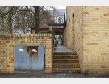 Ein schmaler Durchgang mit Treppen zwischen gelblichen Backsteinmauern, flankiert von einem Baum links. Im Vordergrund befinden sich ein metallisches, leicht verrostetes Stromkastenpanel und eine niedrige Backsteinmauer.