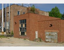 Backsteingebäude mit rechteckigen Formen und großen Metallrohren an der Seite, einer großen Fensterfront und einer verwitterten Metalltür. Der Boden davor ist unbefestigt, und im Hintergrund sind Bäume sichtbar.