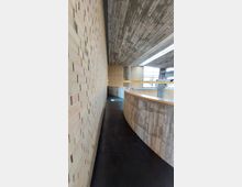 Ein geschwungener Innenraum mit roher Betonarchitektur und Backsteinwänden, einem dunklen Bodenbelag und großen Fenstern, die Tageslicht einlassen. Der Raum hat eine moderne, minimalistische Gestaltung mit sichtbaren strukturellen Elementen.