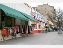 Straßenszene mit Geschäften, darunter ein Bekleidungsgeschäft mit grünen Markisen und Kleidung auf Kleiderständern sowie ein Lebensmittelgeschäft mit Obst- und Gemüseständen im Freien. Im Hintergrund sind mehrstöckige Wohnhäuser und ein baumbestandener Gehweg sichtbar.