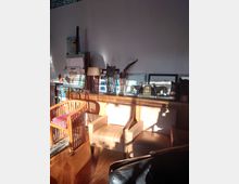 Ein gemütlicher Innenraum mit Holzmöbeln, darunter zwei beige Polstersessel und ein Holzstuhl mit buntem Kissen. An der Wand stehen dekorative Skulpturen, Bilderrahmen und gemalte Leinwände, beleuchtet von einfallendem Sonnenlicht.