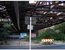 Eine rustikale, metallene Brückenkonstruktion überspannt einen belebten Gehweg, darunter sind Graffiti, Werbetafeln und ein Weg mit Pflanzenbewuchs zu sehen. Im Vordergrund steht ein Verkehrsschild neben einer Straße, während eine Person mit einem Kinderwagen im Hintergrund spaziert.