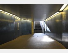 Ein beleuchteter Fußgängertunnel mit dunklen, glänzenden Wänden und einer Treppe am Ende, die ins Tageslicht führt. Beleuchtungskörper sind an der Decke angebracht.