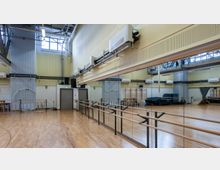 Großer Trainingsraum mit Holzfußboden, Spiegelwand und Ballettstangen. Im Raum befinden sich sportliche Ausstattungen wie Gymnastikreifen, Matten und Leitern, sowie eine hohe Decke mit technischen Installationen und Oberlichtern.