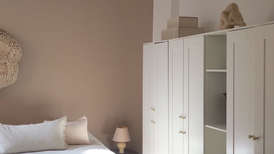Minimalist bedroom with a taupe accent wall, white bed linens, and a large round paper pendant light hanging from a high ceiling with molding. Along one wall are tall white wardrobes with paneled doors and open shelving, set on warm wooden floors beside a small bedside table and lamp.